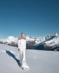 Model in a white off-the-shoulder wedding dress walking on a snowfield with snow-covered mountains under a clear blue sky