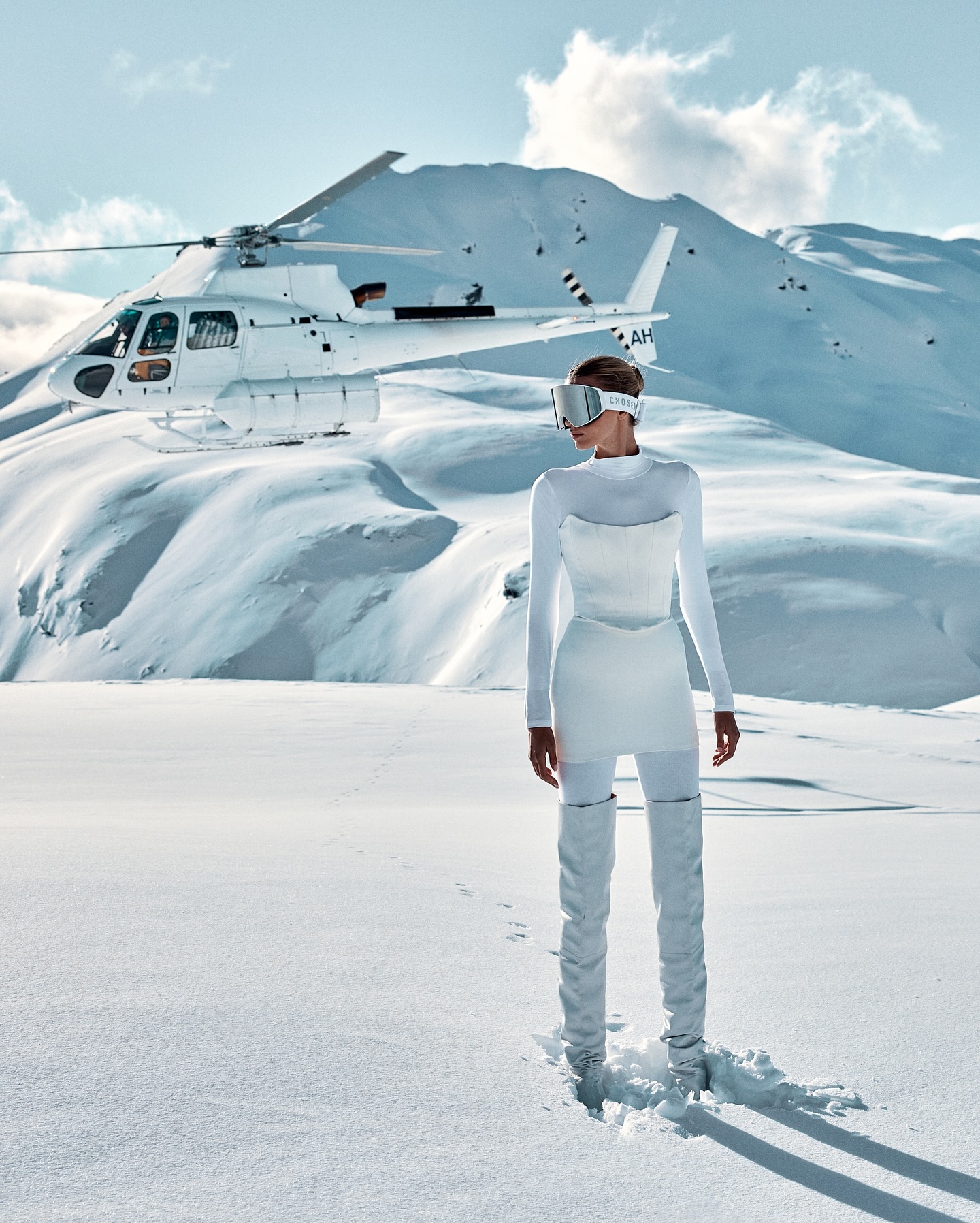 A woman in a white outfit and visor stands in a snowy mountain landscape, with a white helicopter parked on the snow behind her.