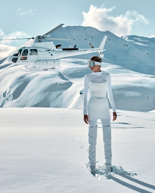 A woman in a white outfit and visor stands in a snowy mountain landscape, with a white helicopter parked on the snow behind her.
