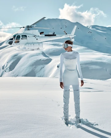 A woman in a white outfit and visor stands in a snowy mountain landscape, with a white helicopter parked on the snow behind her.
