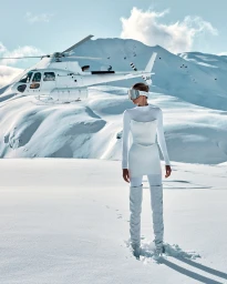 Model in white ski outfit and goggles standing on a snowy mountain with a white helicopter hovering behind.
