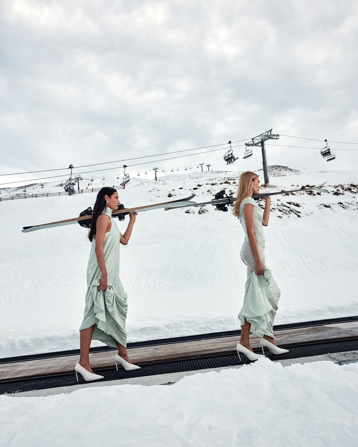 Two women in dresses carry skis in a snowy mountain landscape with a ski lift. The blonde woman is in the foreground.