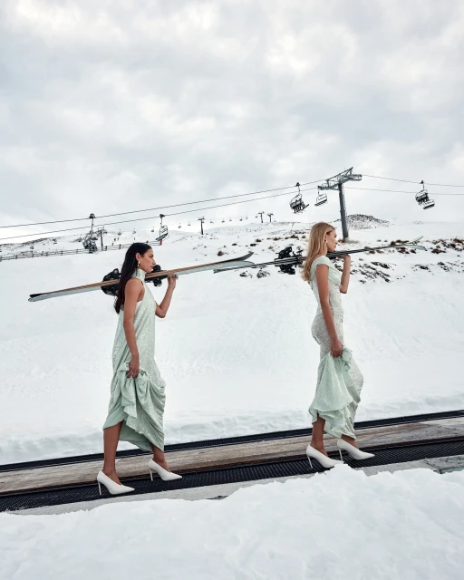 Two women in dresses carry skis in a snowy mountain landscape with a ski lift. The blonde woman is in the foreground.