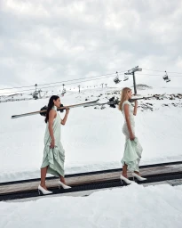 Two models in light green dresses and high heels carrying skis on a snowy mountain with ski lifts in the background.