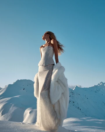 Fashion shoot of a woman in a gown and fur coat standing in a snowy mountain landscape, looking up towards a clear blue sky.
