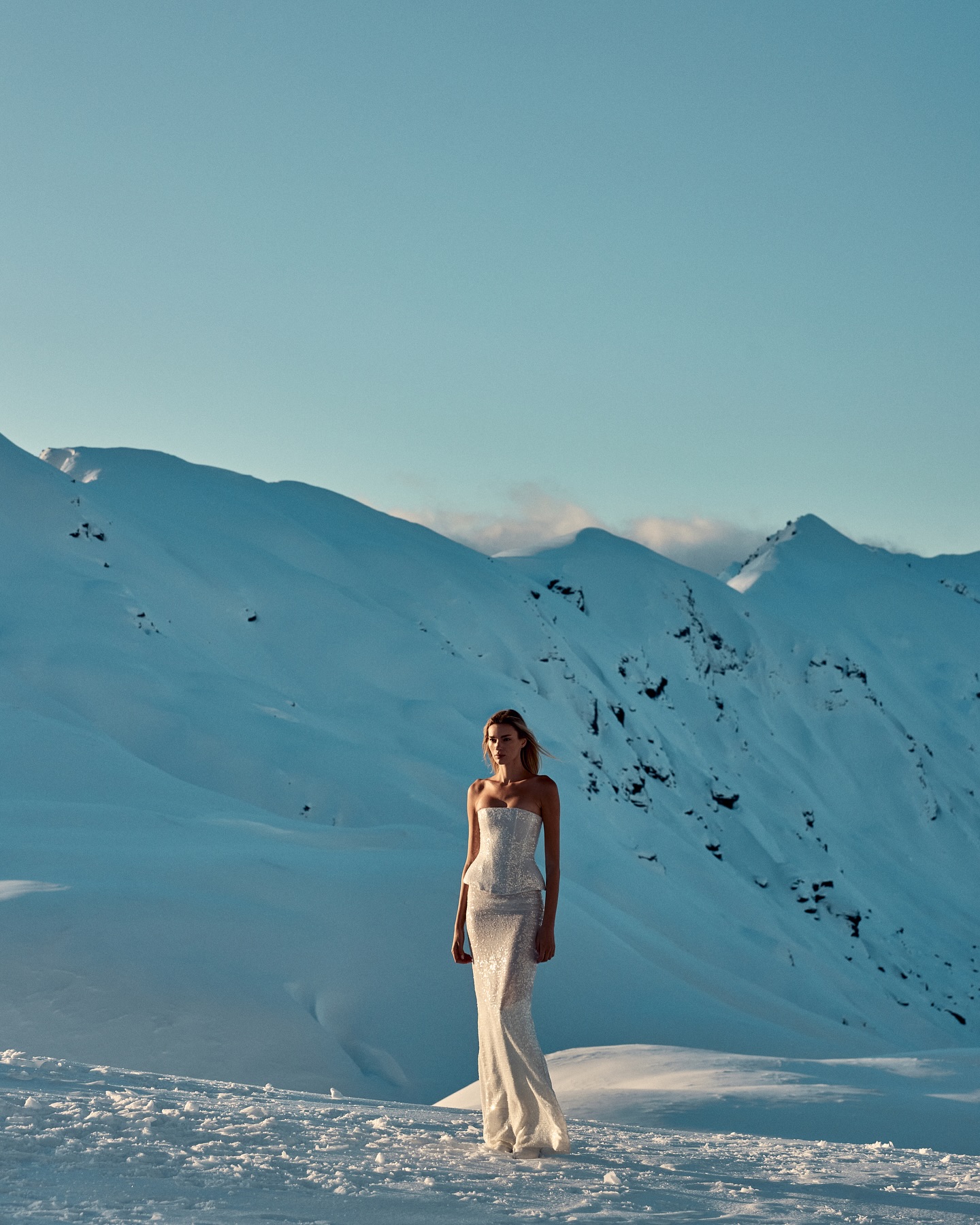 Model standing on a snowy mountain ridge wearing a strapless white gown against a vast alpine landscape