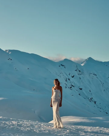 Model standing on a snowy mountain ridge wearing a strapless white gown against a vast alpine landscape