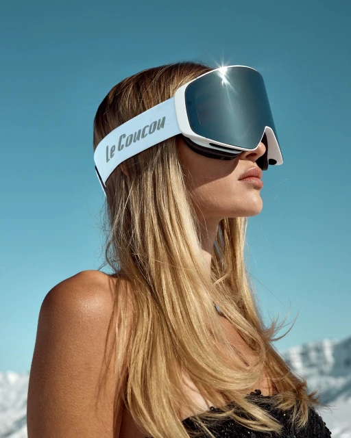A woman with long blonde hair, wearing white Le Coucou ski goggles, poses outdoors against a blue sky and snowy mountain background.