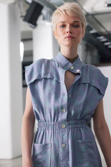 Woman with short blonde hair wearing a blue, button-down dress with pockets in a bright, minimalist studio environment.