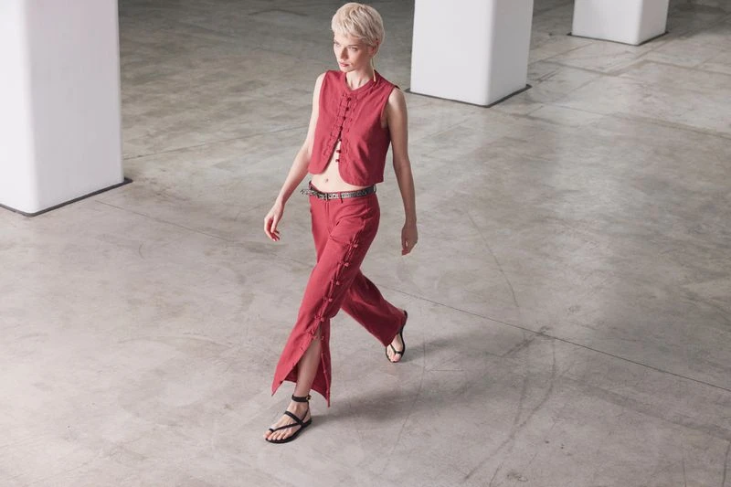 A woman strides confidently in a red sleeveless top and matching pants, complemented by black sandals, against a stark, grey backdrop.