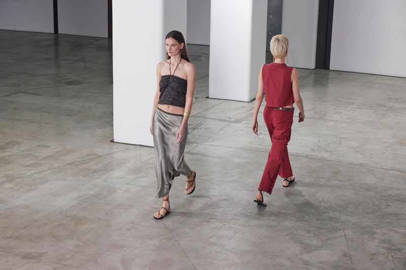 Two women walk in a fashion show, wearing a black halter top and silver skirt, and a red vest and matching trousers, on a gray floor.