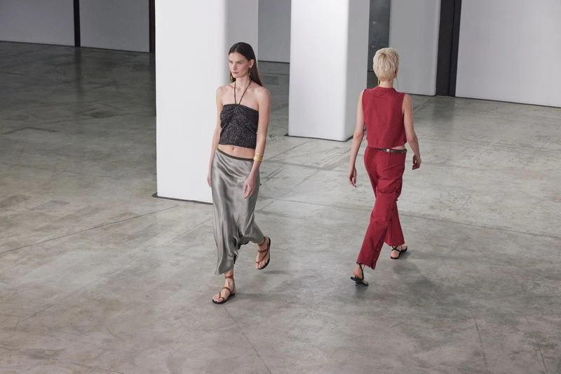 Two women walk in a fashion show, wearing a black halter top and silver skirt, and a red vest and matching trousers, on a gray floor.
