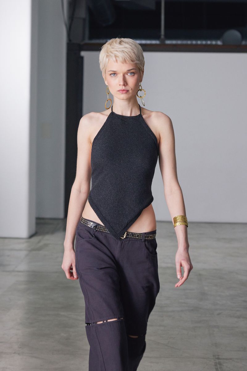 A woman in a black halter top and loose black pants poses indoors in a fashion setting with neutral-toned floors and a backdrop.