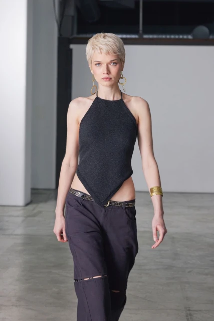 A woman in a black halter top and loose black pants poses indoors in a fashion setting with neutral-toned floors and a backdrop.