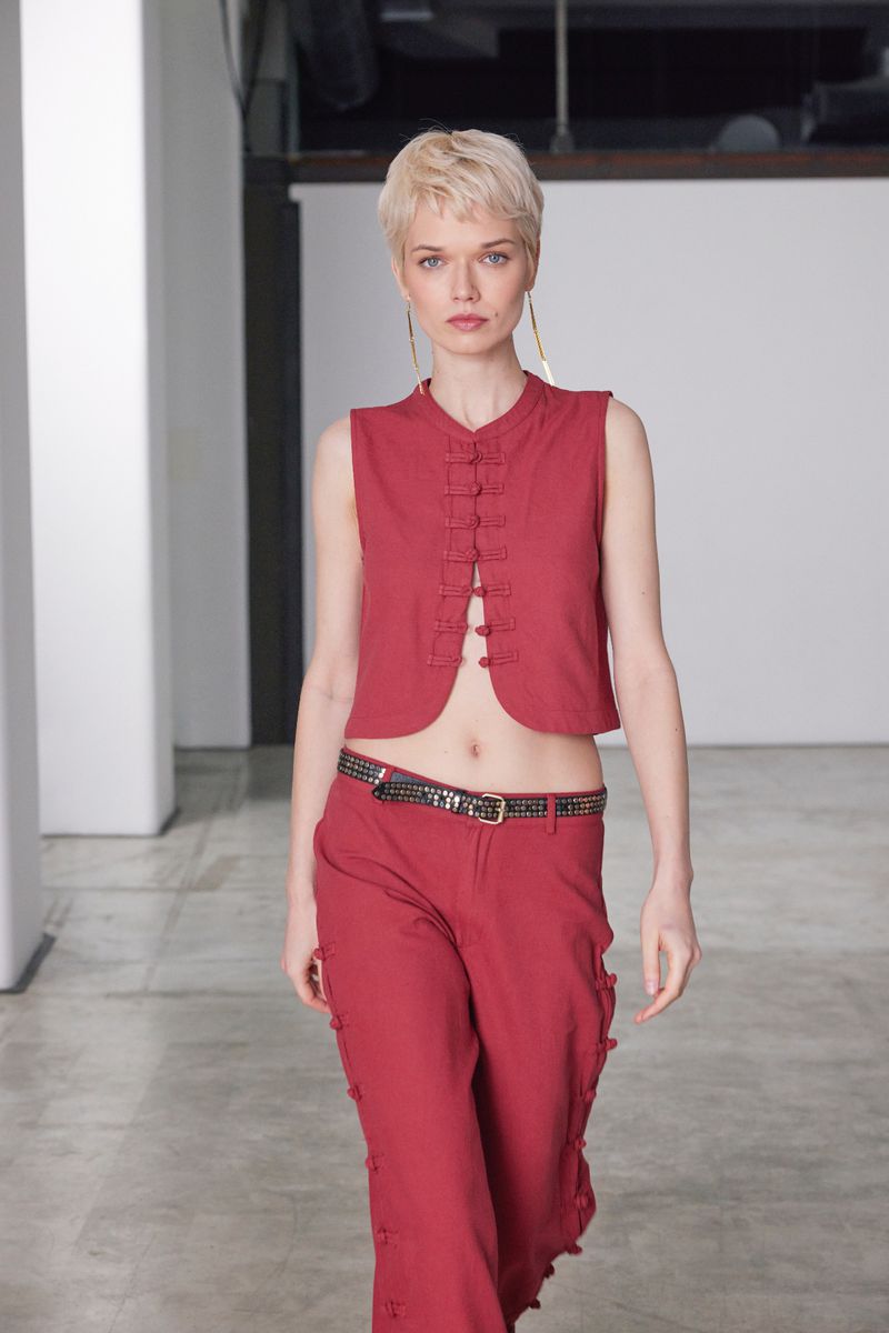 Fashion model in red vest and pants walks on a concrete floor with a white wall and large windows in the background in a photo studio.