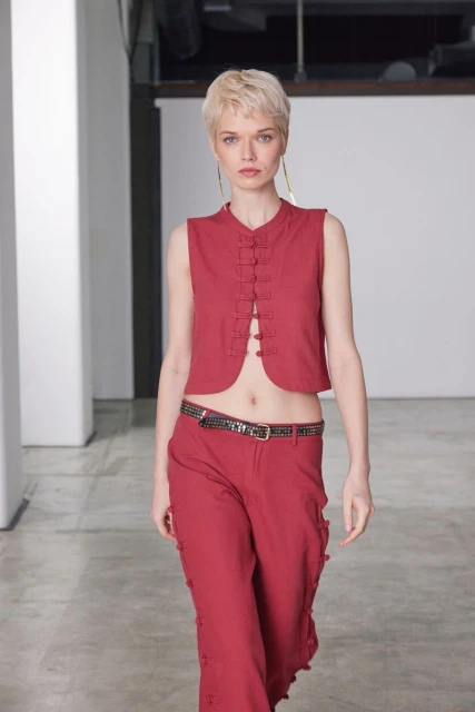 Fashion model in red vest and pants walks on a concrete floor with a white wall and large windows in the background in a photo studio.