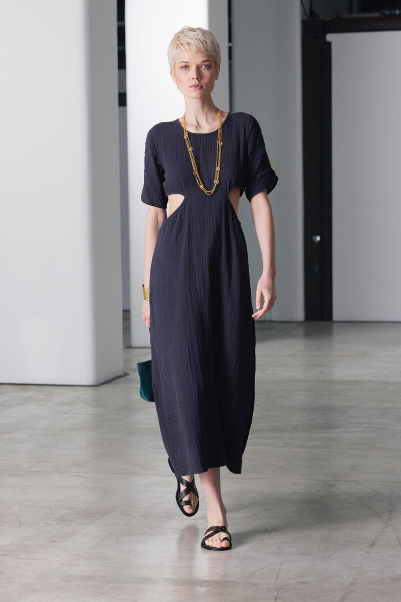 A woman with short blonde hair in a navy midi dress and black sandals walks on a light grey studio floor with white background.