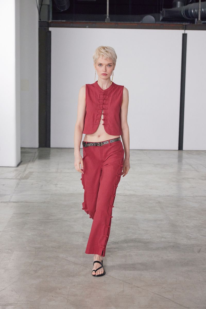 Fashion model posing in a red sleeveless top and pants in a studio setting, with a concrete floor and industrial background.