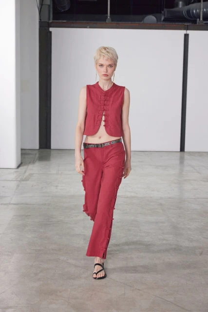 Fashion model posing in a red sleeveless top and pants in a studio setting, with a concrete floor and industrial background.