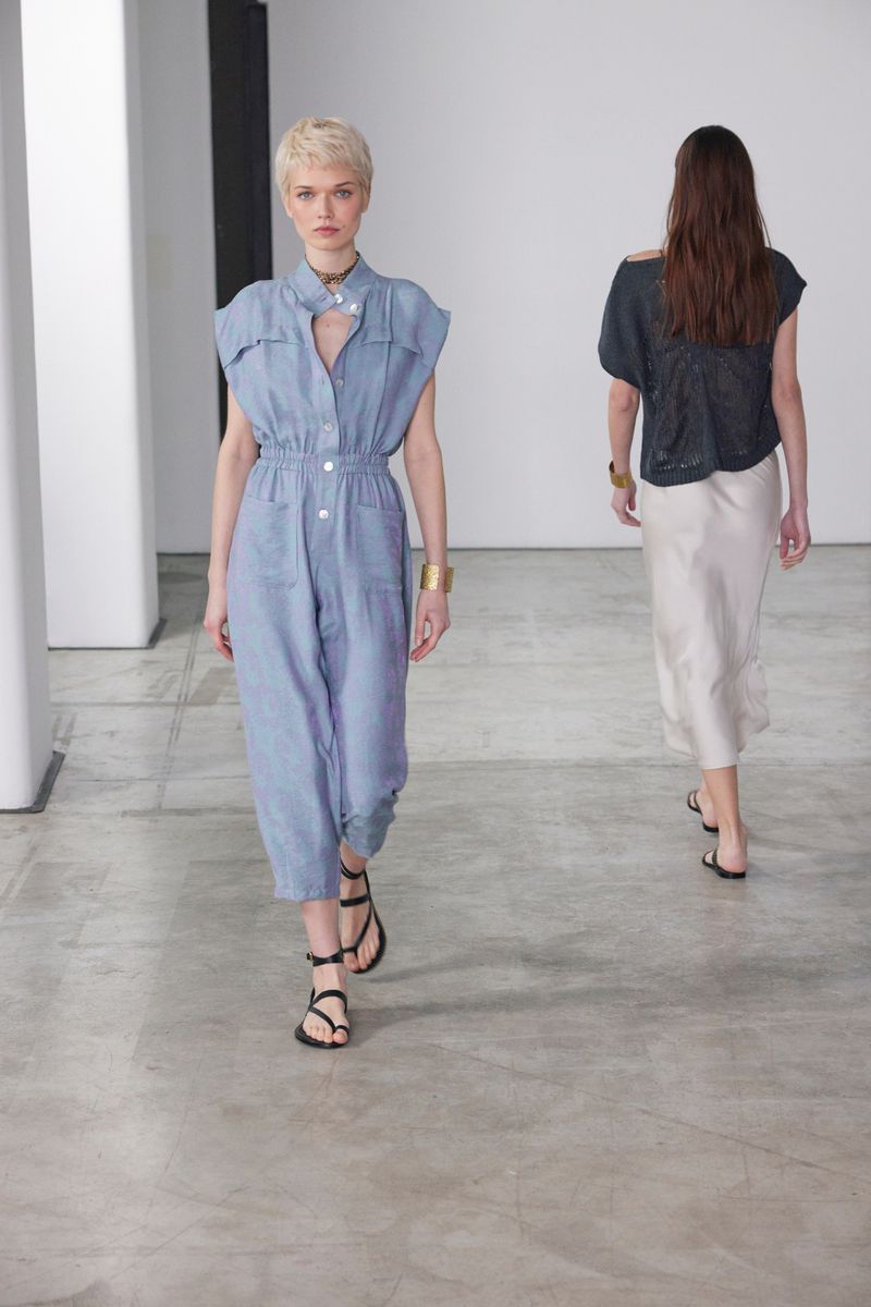 Two fashion models on a concrete runway. The foreground model wears a blue jumpsuit and sandals. The background model wears a black top and skirt.