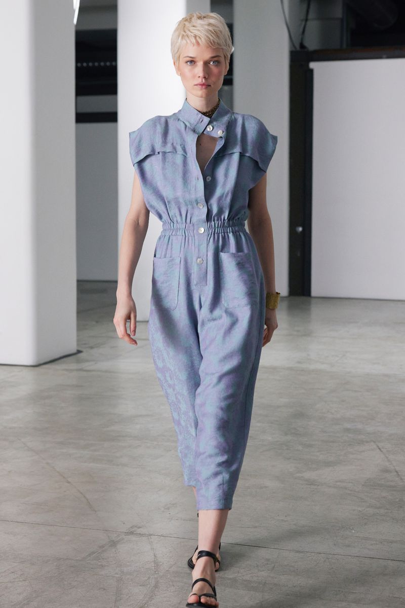 Woman in blue jumpsuit and black sandals walking on a concrete floor in a bright studio setting. Fashion editorial or runway shot.