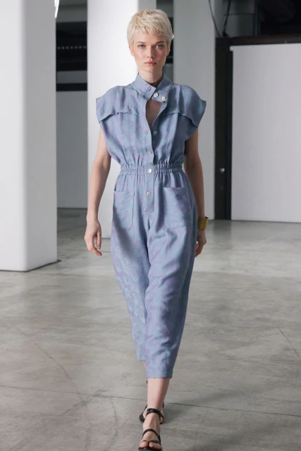 Woman in blue jumpsuit and black sandals walking on a concrete floor in a bright studio setting. Fashion editorial or runway shot.