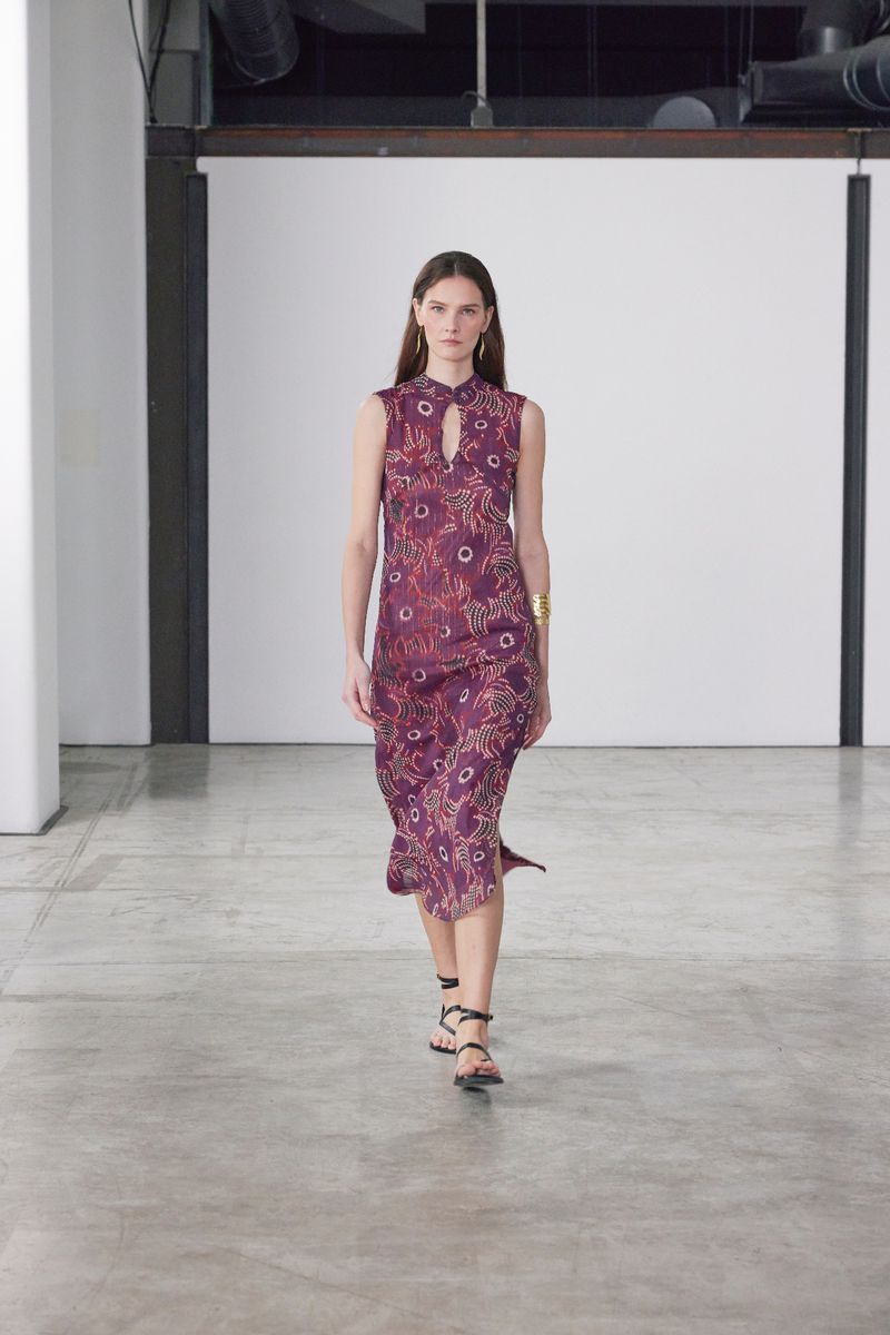 A woman in a patterned sleeveless dress walks on a gray concrete floor in a minimalist indoor setting, accessorized with gold bracelets.