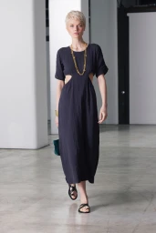 Model with short blonde hair wearing a black dress with side cutouts and gold layered necklaces walking indoors
