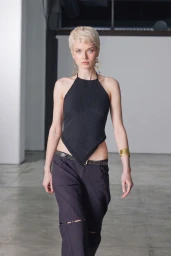 Short-haired blonde model walking wearing a black halter top and dark pants against an indoor studio background
