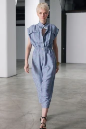 Model with short blonde hair wearing a blue button-down jumpsuit walking in a modern architectural interior space