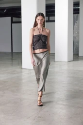 Model walking forward in a shiny gray midi skirt and a halter neck top inside a spacious modern hall with columns.