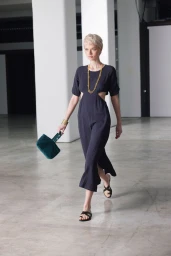 Model with short blonde hair wearing a black cutout dress and holding a green handbag walking across a polished concrete floor