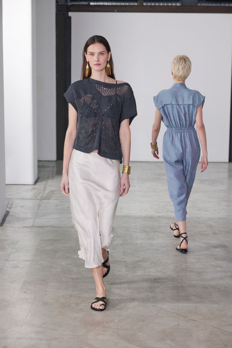 Two female models walking in a bright indoor studio space wearing modern clothing including a dark grey knit top and a light skirt