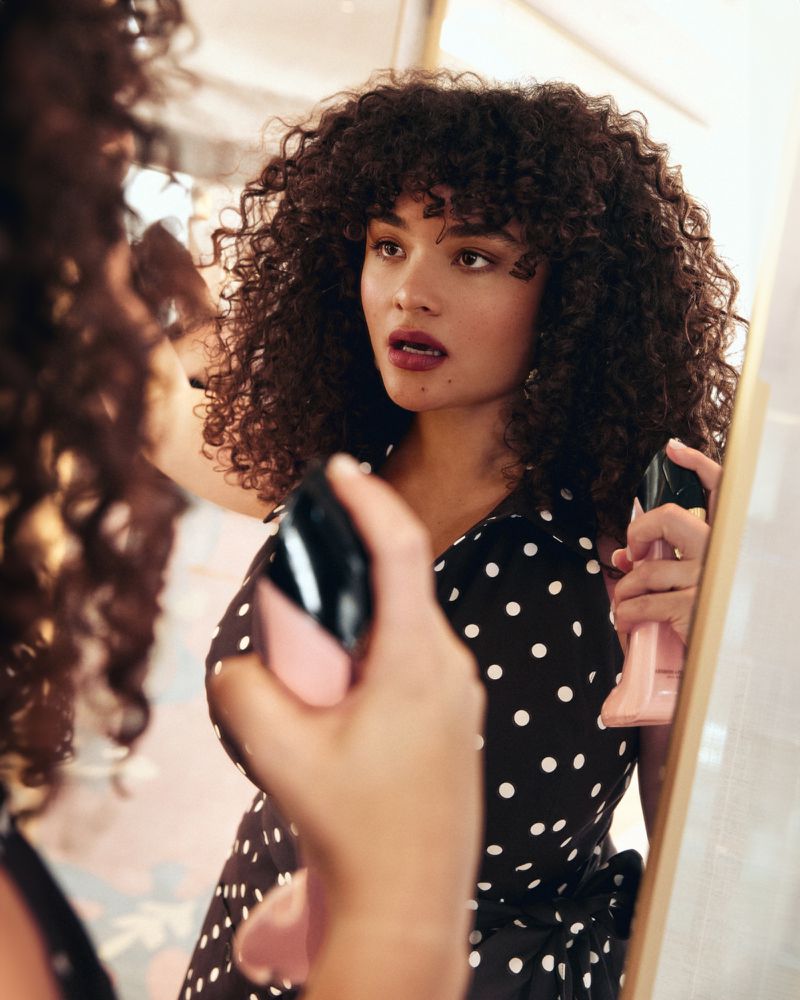 Woman with curly hair in polka dot dress applies makeup in a mirror; beauty shot in an indoor environment.
