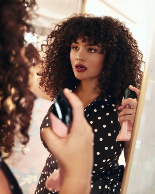 Woman with curly hair in polka dot dress applies makeup in a mirror; beauty shot in an indoor environment.