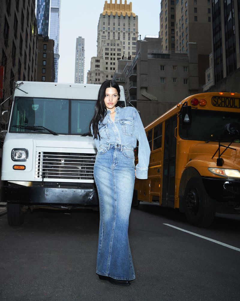 A woman wearing a denim jacket and skirt stands in a street with a white truck and yellow school bus in the background, buildings behind.