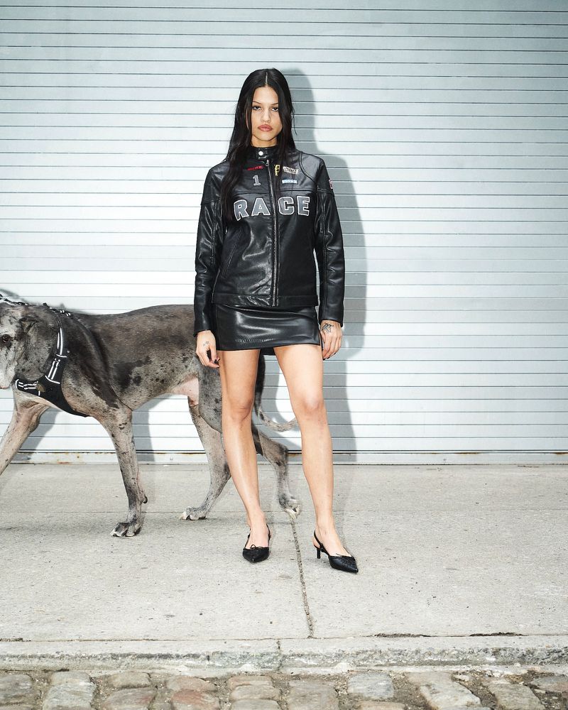 Woman in a leather jacket and skirt poses with a large dog against a metal garage door and cobblestone. Fashion editorial shot.