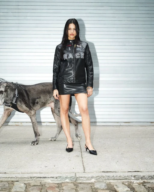 Woman in a leather jacket and skirt poses with a large dog against a metal garage door and cobblestone. Fashion editorial shot.