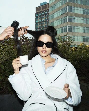 Adult woman in bathrobe and sunglasses sits outdoors drinking coffee while her hair is styled; buildings in the background.