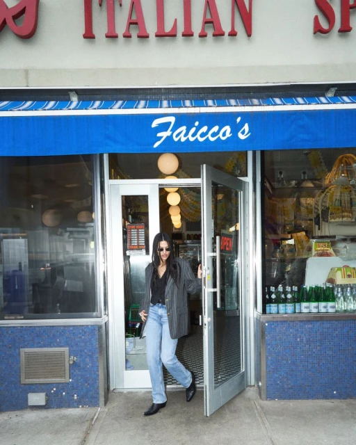 Woman exits Faicco's Italian Specialties in New York City, wearing a blazer, light-wash jeans, and sunglasses, captured in a medium shot.