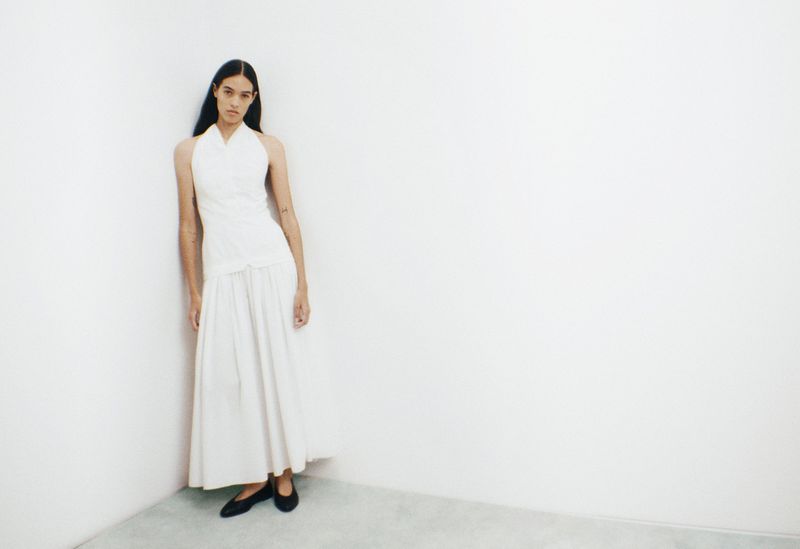 A slender adult woman in a white dress leans against a white wall, with dark hair, wearing black ballet flats in an indoor space.