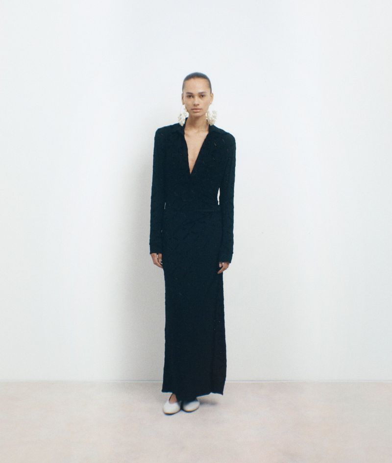 Elegant woman posing in long-sleeved, floor-length black dress, adorned with large ornate earrings, against a white backdrop in studio.