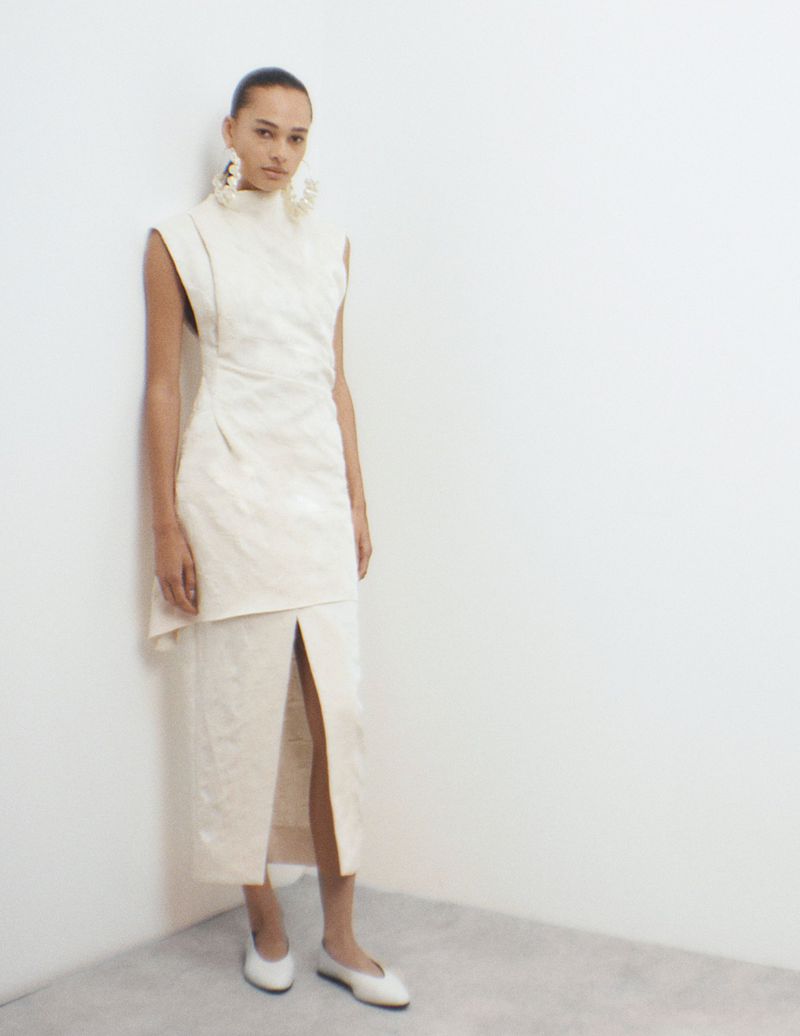 A woman poses in a cream-colored high-necked dress with a thigh-high slit and white flats against a neutral corner backdrop, studio lighting.