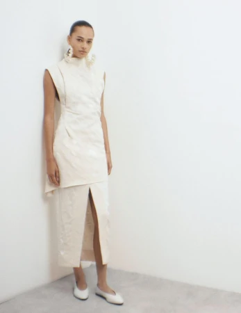 A woman poses in a cream-colored high-necked dress with a thigh-high slit and white flats against a neutral corner backdrop, studio lighting.