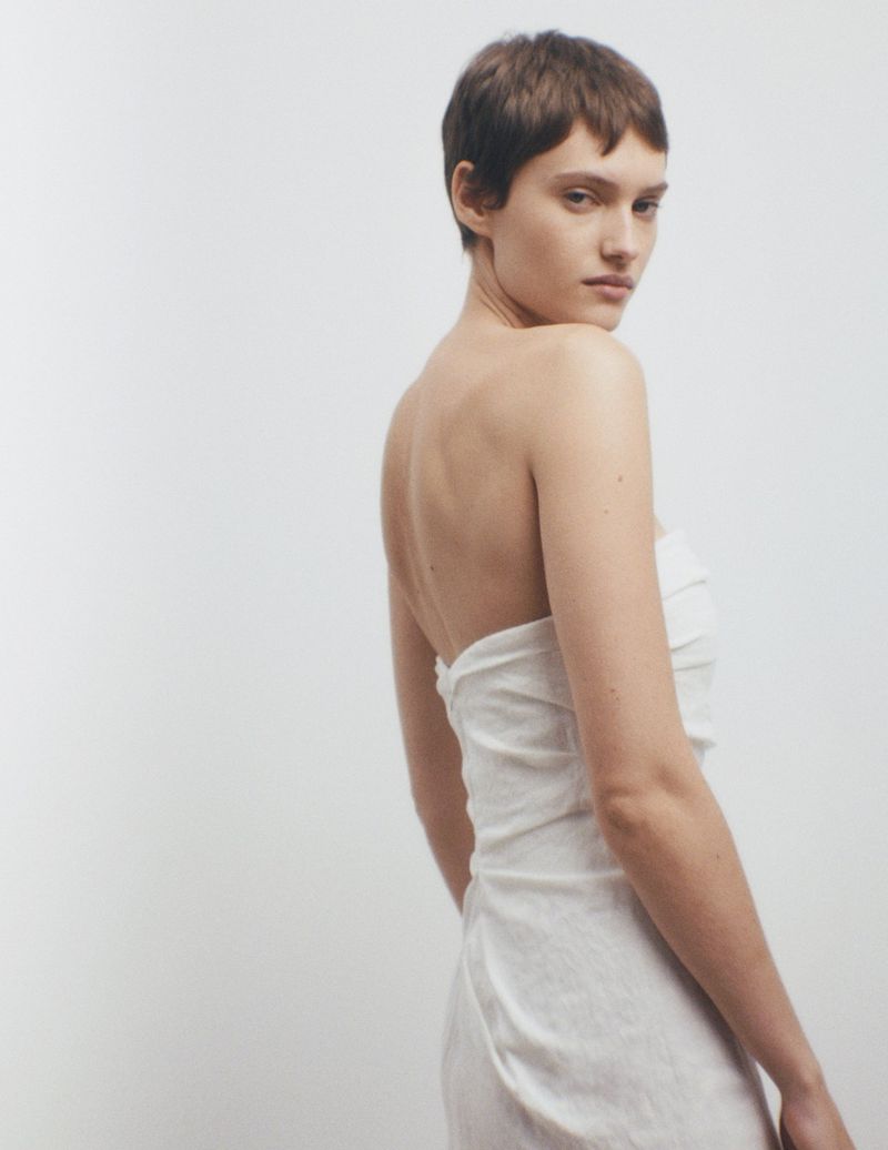 Woman with short brown hair poses in a white strapless dress against a clean white backdrop, looking back over her shoulder.