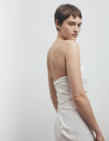 Woman with short brown hair poses in a white strapless dress against a clean white backdrop, looking back over her shoulder.