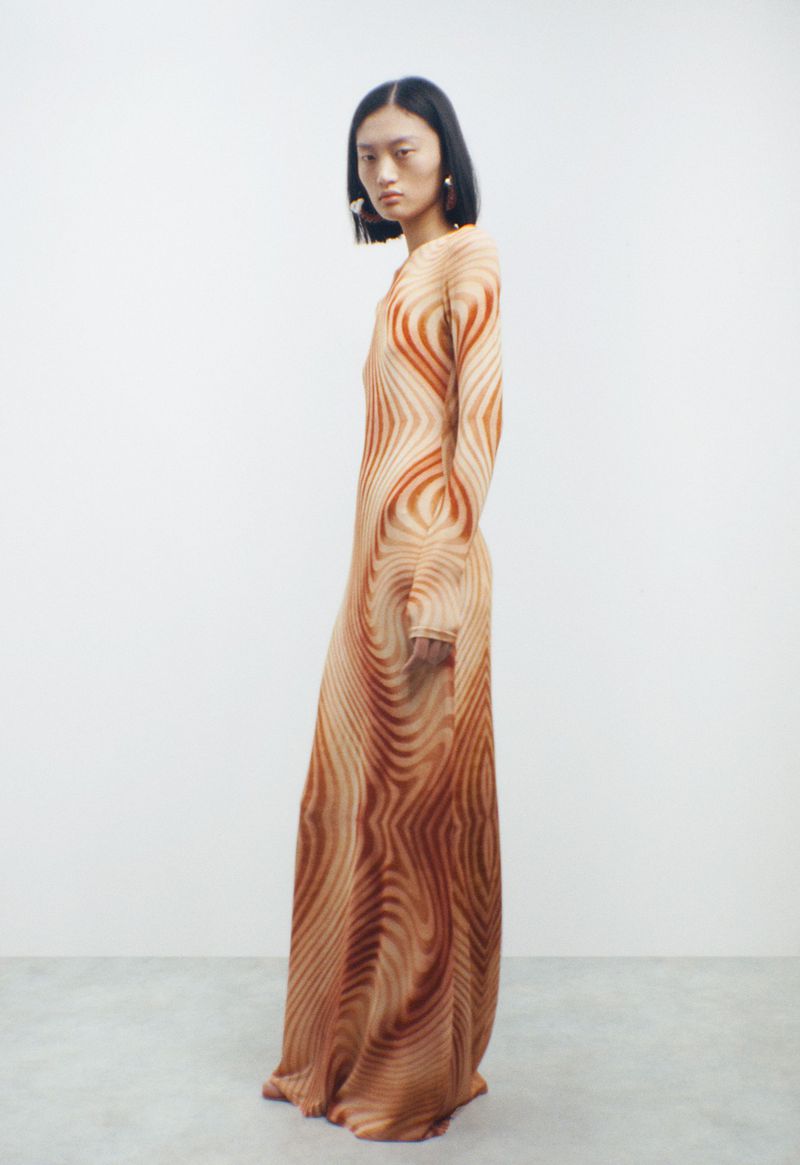 Asian female model posing in a long-sleeve gown with a swirly pattern, standing in a bright studio with a neutral facial expression.