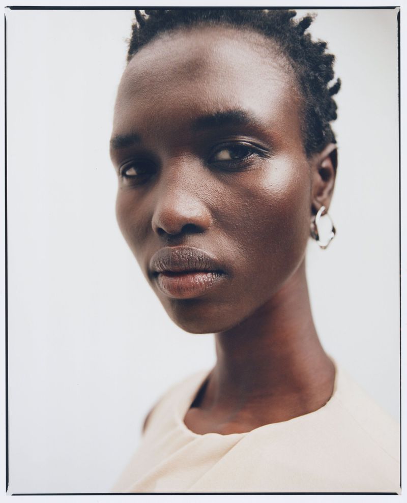 Close-up portrait of a dark-skinned woman with short dark hair wearing a beige top and hoop earrings against a plain white background.