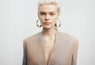 Portrait of a model with short blonde hair wearing sculptural gold earrings and a structured blazer against a plain white background.