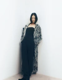 Model wearing a black jumpsuit and zebra-print coat leaning against a white corner wall